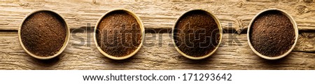 Four varieties of freshly ground coffee powder in individual dishes showing medium and full roast on a cracked weathered driftwood background, overhead view in banner format