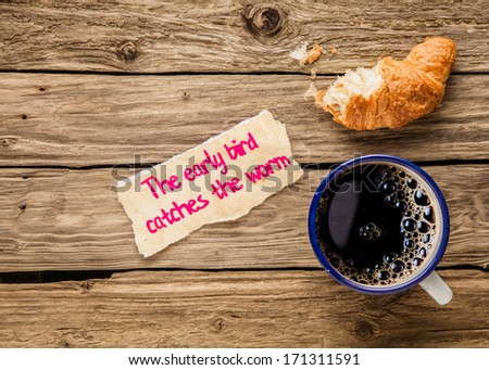 The early bird catches the worm, an inspirational saying hand written on a small torn piece of paper alongside an early breakfast of frothy espresso coffee and a half devoured fresh golden croissant