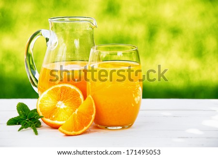 A jug and glass of freshly squeezed orange juice, garnished with a sprig of mint and slices of orange, in an outdoor setting during summer.