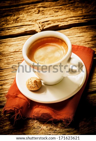 Cup of hot frothy espresso coffee served on a rustic cloth with a small macaroon in a rural country cafeteria on a weathered wood surface with vignetting and copyspace