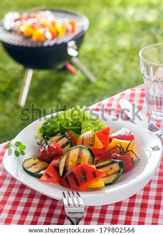 Delicious healthy plate of colorful roasted vegetables with marrow, tomato , sweet peppers and herbs garnished with frilly lettuce on a picnic table in a summer garden with a barbecue behind