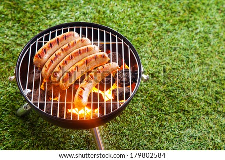 Sausages grilling over the hot glowing coals in a portable barbecue standing on a green lawn with copyspace during a picnic or summer camping trip