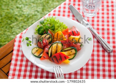 Healthy country roasted vegetables, vegetarian food, served in a bowl on a red and white checked tablecloth on a picnic table outdoors with peppers, marrow, herbs and tomato