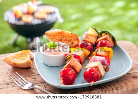 Healthy summer meal of halloumi and vegetable kebabs roasted over an outdoor barbeque in the garden and served with a savory sauce and toasted baguette on a picnic table