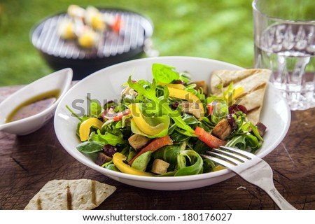 Tasty herb salad with fresh green leaves of rocket, sweet pepper and tomato served with an oil dressing on a summer picnic table in the garden