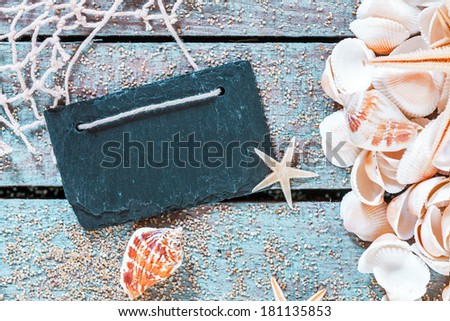 Pile of assorted seashells with a blank retro school slate with copyspace on rustic wooden table with scattered beach sand, fishing net and starfish