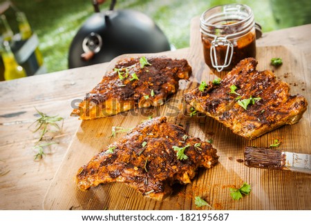 Basting and seasoning ribs for the BBQ with three portions of meat laid out on a wooden picnic table in a summer garden with a jar of basting sauce and herbs