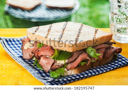 Grilled toasted sandwich with rare roast beef, rocket and herbs on a blue and white checked cloth outdoors on a picnic table at a summer BBQ