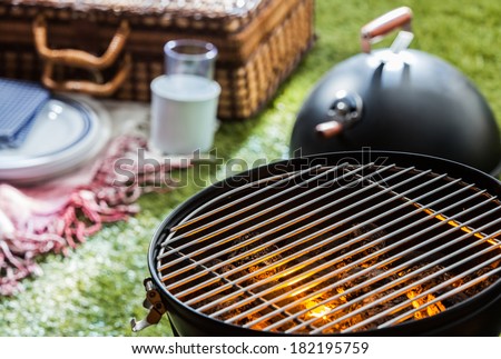 Close up of a burning hot fire in a portable barbecue with an empty grill and a wicker picnic hamper visible on a green lawn behind