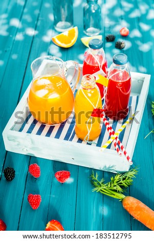 Bottles of freshly squeezed homemade citrus and berry juice with a jug of iced carrot and orange juice blend served on a wooden tray on a colorful turquoise picnic table outdoors