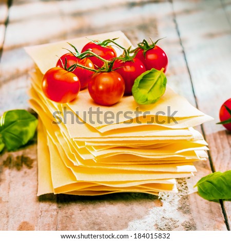 Dried uncooked lasagne pasta sheets with fresh ingredients including cherry tomatoes and basil leaves, close up on old wooden boards