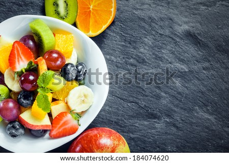 Overhead view of a bowl of fresh fruit salad for a healthy diet made with assorted tropical fruits and an apple, kiwifruit and orange arranged alongside on textured slate with copyspace