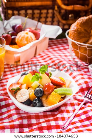 Healthy vegetarian summer picnic with a box of assorted whole fresh fruit and bowl of colourful tropical fruit salad with a hamper on a red and white checked tablecloth