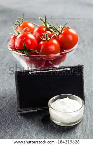 Glass bowl of fresh ripe red cherry tomatoes and savory creamy vinaigrette sauce on a slate background with a blank vintage school slate with copyspace for your text