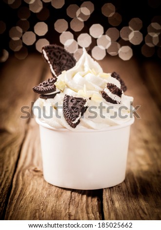 Pieces of broken chocolate oreos on a twirl of rich creamy frozen yoghurt in a plastic takeaway tub on a wooden counter with a bokeh of twinkling lights