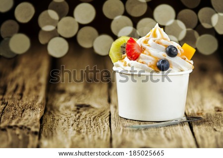 Creamy vanilla frozen yogurt topped with fresh tropical fruit served in a plastic takeaway tub with a disposable teaspoon on a wooden table with a background bokeh of party lights