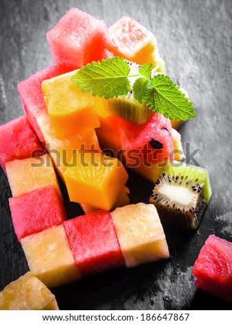 Pile of diced and cubed fresh summer fruit being prepared for a gourmet dessert with watermelon, pineapple, melon, orange, and kiwifruit on a slate kitchen counter