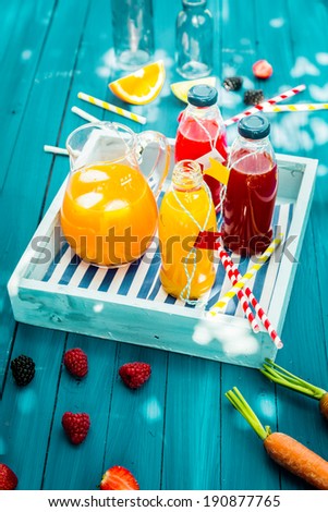 Fresh healthy homemade fruit juice squeezed from citrus, raspberries strawberries and carrots standing on a wooden tray in glass bottle on a turquoise picnic table in dappled summer sunlight