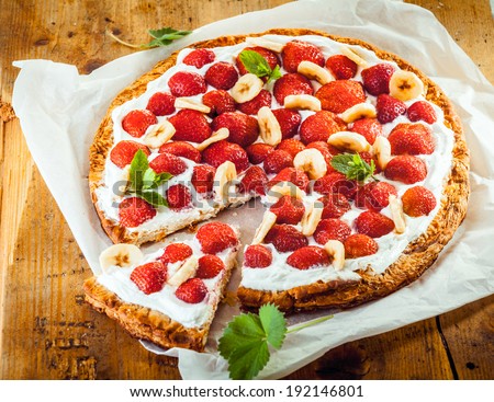 Freshly baked strawberry and banana flan with a crisp golden crust and topping of fresh berries , banana and cream garnished with peppermint on a square of white paper, high angle view
