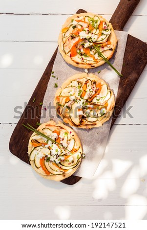 Three cooked delicious mini vegetarian pizzas on a long wooden board standing on a white painted wooden table outdoors in dappled sunlight, overhead view