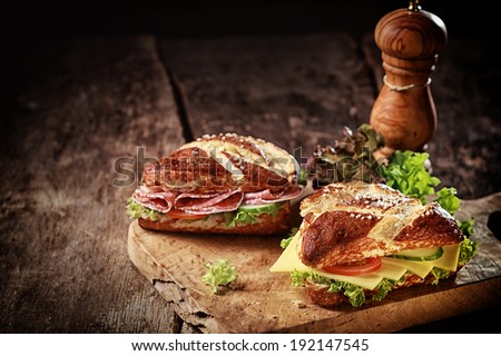 Crusty brown lye bread roll sandwiches with a cheese, salami, lettuce, tomato and cucumber filling on an old grunge board with a wooden pepper mill and copyspace