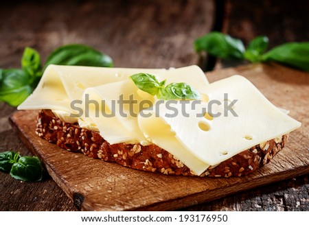 Thinly sliced gouda cheese on wholewheat bread garnished with fresh basil served on a wooden board, close up view