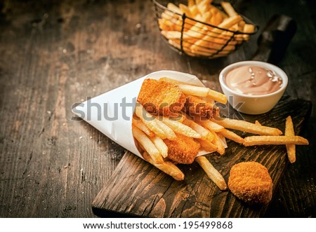 Serving in a paper cone of takeaway crumbed fried fish nuggets with potato chips and a small bowl of sauce or dip on a rustic wooden table with copyspace