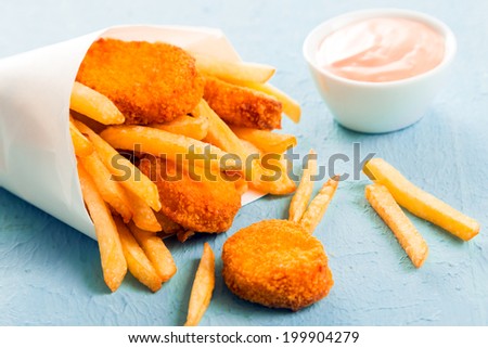 Fried fish nuggets with golden French fries spilling out of a paper cone from a takeaway fish shop onto a blue surface, close up view