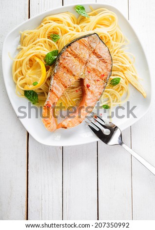 Gourmet seafood cuisine with grilled salmon cutlet steak served on a bed of Italian linguine pasta garnished with lemon and fresh basil, overhead view on a white wooden table with copyspace