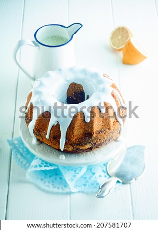 Freshly baked glazed lemon ring cake served on a cake stand for dessert with a jug of tangy citrus sauce and a spatula to serve, high angle view