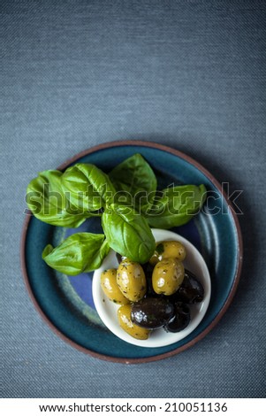 Overhead view of a bunch of fresh basil leaves with a bowl of black and green cured olives on a grey weave textile background with copyspace
