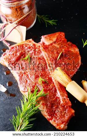 Raw porterhouse steak with spicy seasoning and a sprig of fresh rosemary alongside a jar of the powdered spice with a blank label during preparation of the meat for grilling in a rustic kitchen