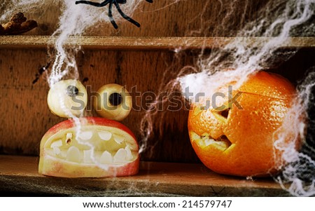 Fun spooky homemade Halloween food decorations with a carved apple mouth full of fearsome teeth topped with round dough eyes and a jack-o-lantern carved orange on a wooden shelf with spiders and webs