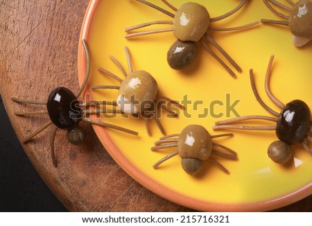 Cute spooky Halloween spider appetizers made from green, black and stuffed olives with spaghetti legs crawling off a yellow platter onto a wooden table, view from above