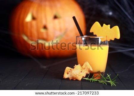 Glass of fresh orange juice decorated for Halloween with a flying bat served with a bat cookie and straw with a pumpkin jack-o-lantern in the background