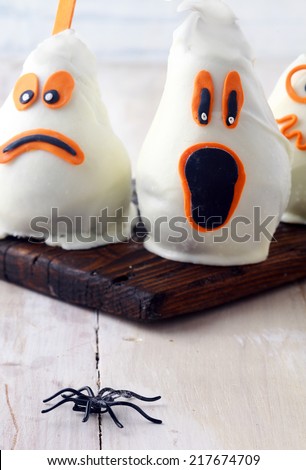 Scary edible Halloween party decorations with fresh pears covered in white chocolate and marzipan to resemble ghosts with spooky expressions on a rustic white table with a spider