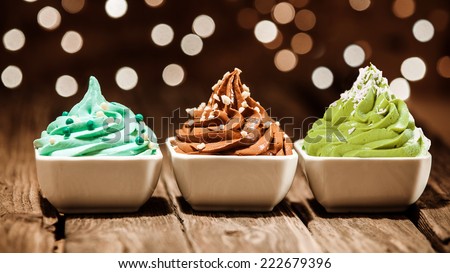 Colorful row of three different frozen yogurt desserts in blue, brown and green garnished with nuts and sugar pearls at a party with a sparkling background bokeh of lights