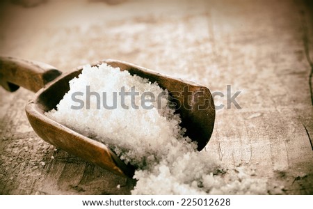 Coarse granules of natural rock or sea salt in a rustic old wooden ladle spilling out onto a grunge wooden table