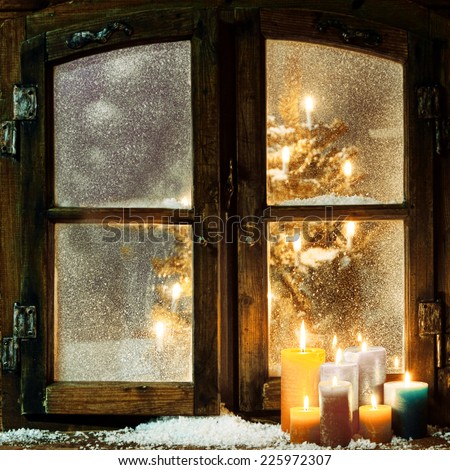 Welcoming Christmas window in a log cabin with a group of burning candles on the windowsill and a glowing Christmas tree visible through the frosted panes