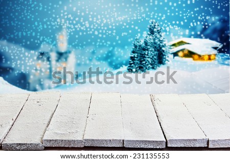 Empty rustic white painted wooden table with rough boards against a winter Christmas scene of a log cabin and church in snowy mountain scenery with glowing windows for product placement