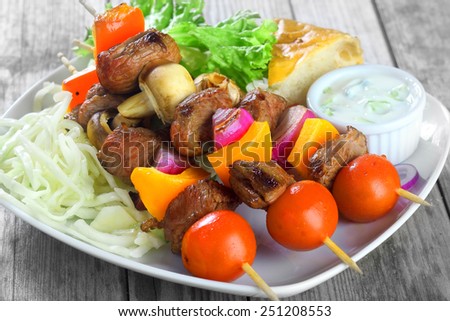 Close up Gourmet Kebabs on White Plate with Mustard Sauce, Fresh Veggies and Bread Slice. Placed on Wooden Table.