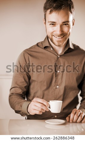 Faded effect image of a smiling happy bearded young man in a brown shirt enjoying a cup of coffee sitting at a table looking at the camera with a beaming grin