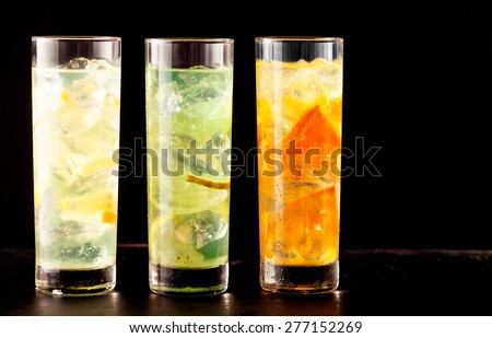 Three highball glasses filled with alcoholic or non-alcoholic cocktail drinks mixed with ice and tropical fresh fruits as lemon, lime and orange, on a table, close-up with copy space on black