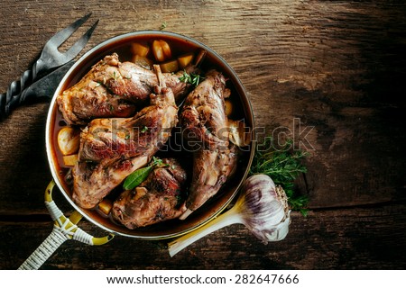 High Angle View of Roasted Rabbit Haunches in Pan with Stewed Vegetables on Rustic Wooden Table Surface with Bulb of Garlic and Copy Space