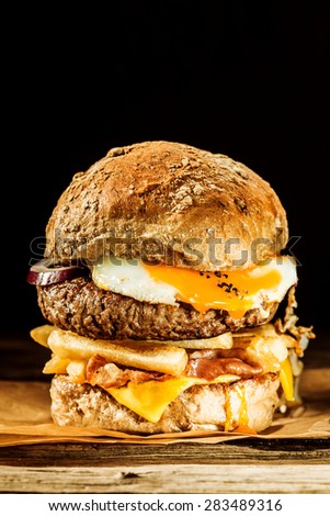 Wholesome egg and bacon burger with a beef patty, fried egg and bacon, cheese, onion and french fries on a wholegrain roll served as a rustic takeaway on brown paper
