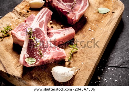 High Angle Close Up View of Raw Lamb Filets Seasoned with Garlic, Salt and Fresh Herbs on Rustic Wooden Board on Dark Textured Surface