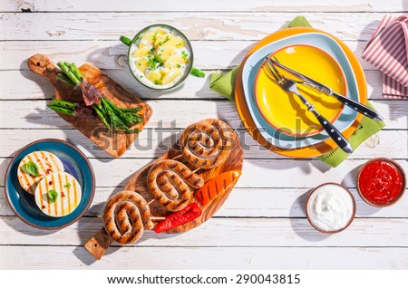 High Angle View of Barbequed Meal of Grilled Sausages and Prepared Side Dishes Arranged on White Picnic Table