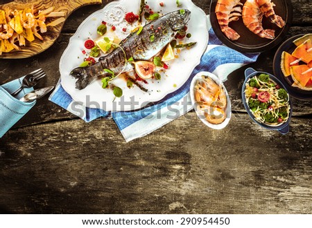 Whole Grilled Fish on White Platter Surrounded by Seafood Dishes on Rustic Wooden Table with Linen Napkins and Cutlery with Copy Space