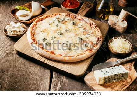Fresh ingredients for a four cheeses Italian pizza laid out in individual bowls on a rustic wooden kitchen counter around a baked pizza with tomato paste and herbs and a melted cheese topping