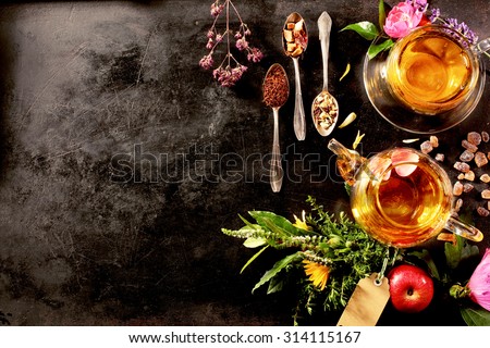 Overhead view of various sorts of tea. Flavoured with Assorted Herbs, an apple, rose with a teapot and a teacup on a rustic black metal board